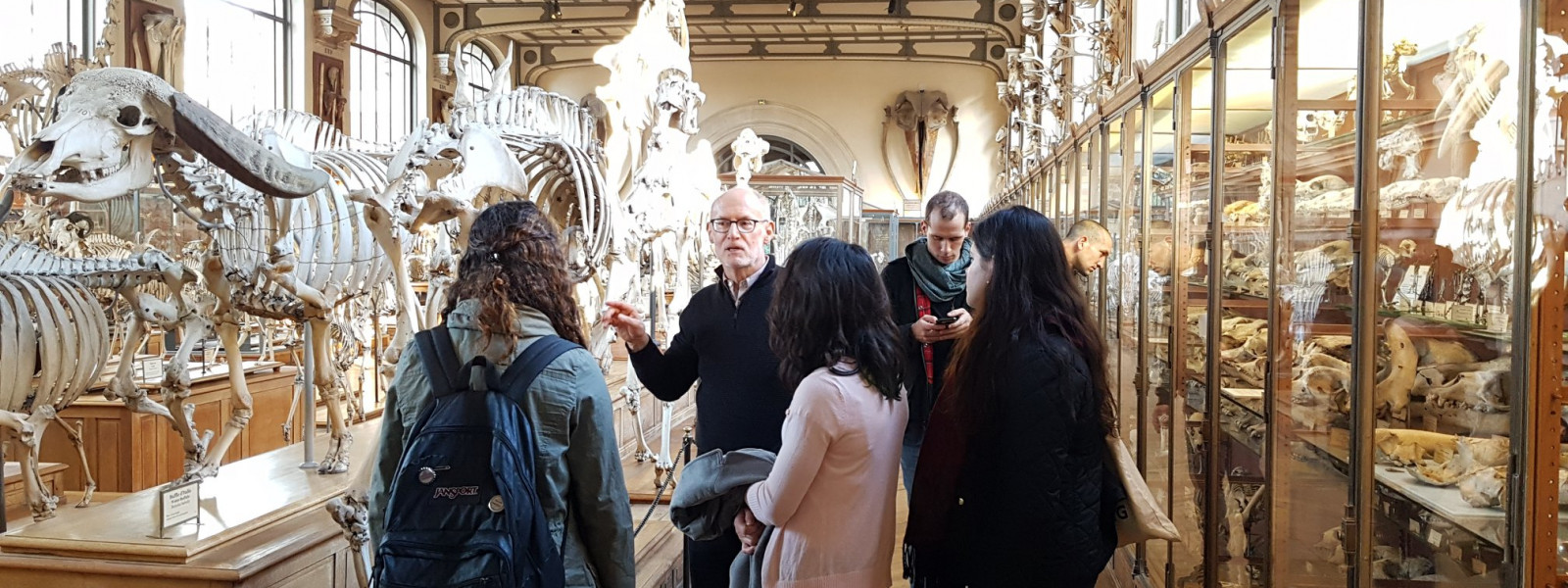 A few students stand listening to the professor lecture by an exhibit of animal skeletons.