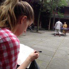 A student is sitting in a courtyard and looking down at a notebook with pen in hand. Two monks play ping pong in the background.