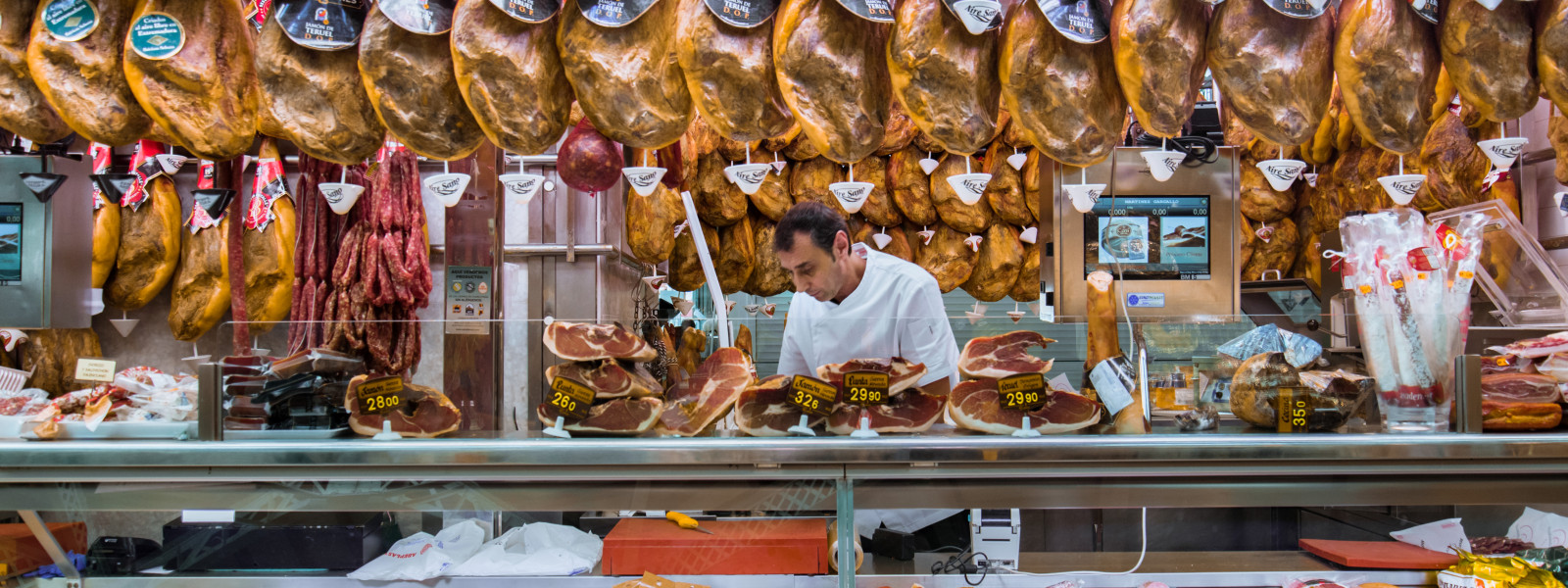 A person behind the counter of a well-stocked jamon shop appears to be immersed in their work