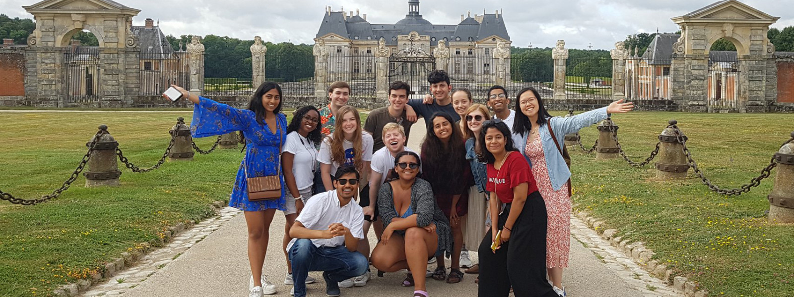 Fifteen students pose on a path in front of the château. The students on either end of the group have their arms spread wide.