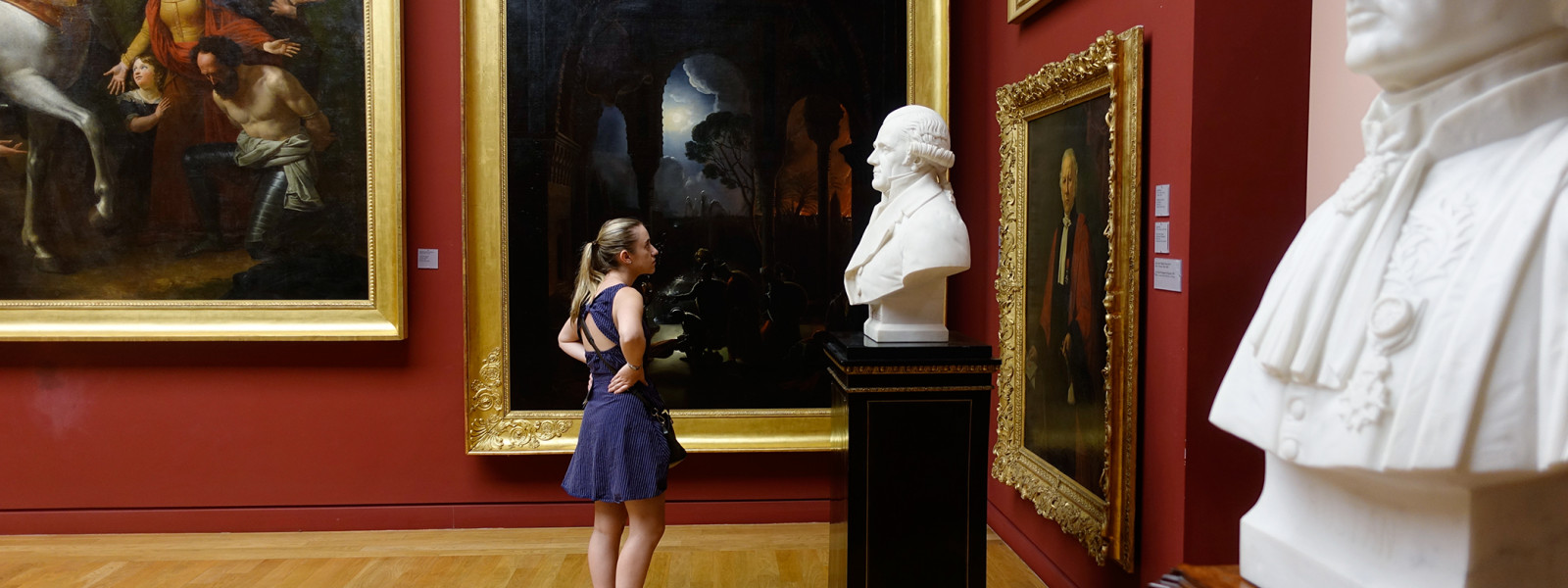 A student stands in a gallery and studies a sculpture while surrounded by paintings.