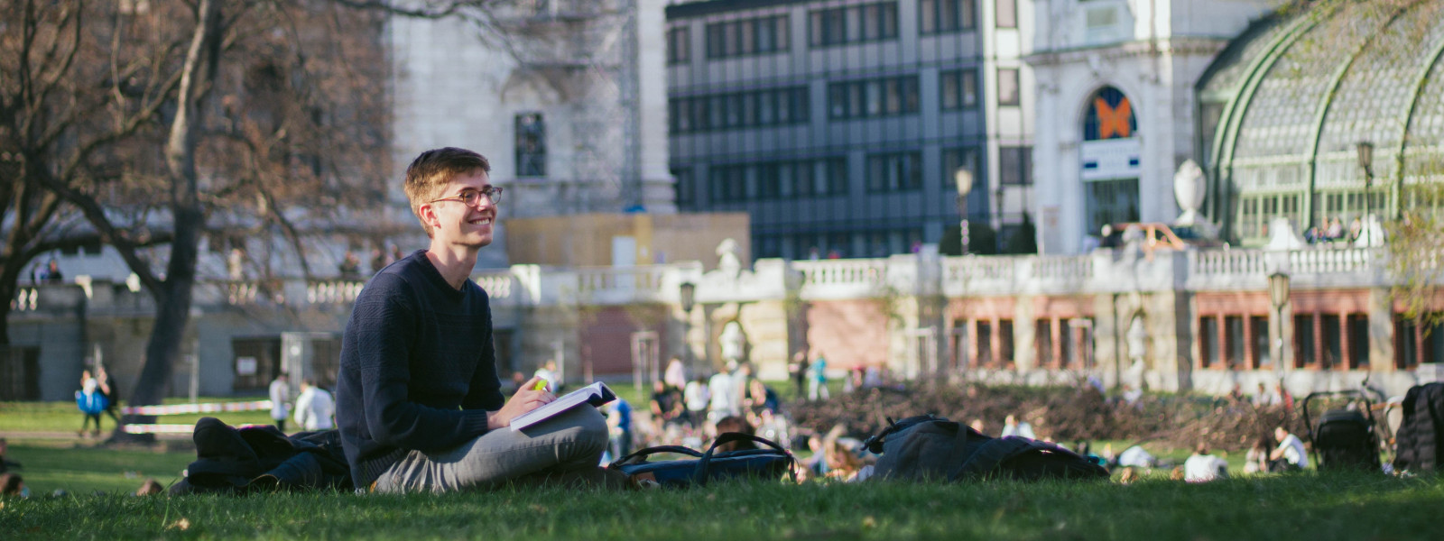 A student sits on the grass of an urban park on a sunny day and looks up from a book, smiling.