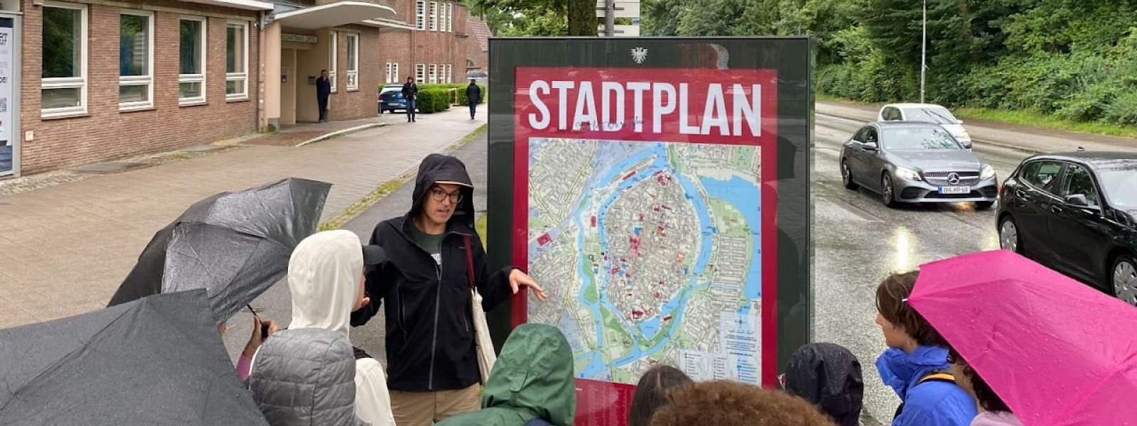 The group gathers outdoors around the professor, who is speaking while standing next to a map. Many of the participants are holding umbrellas. Behind them are a large brick building, trees, and a road with cars passing by.