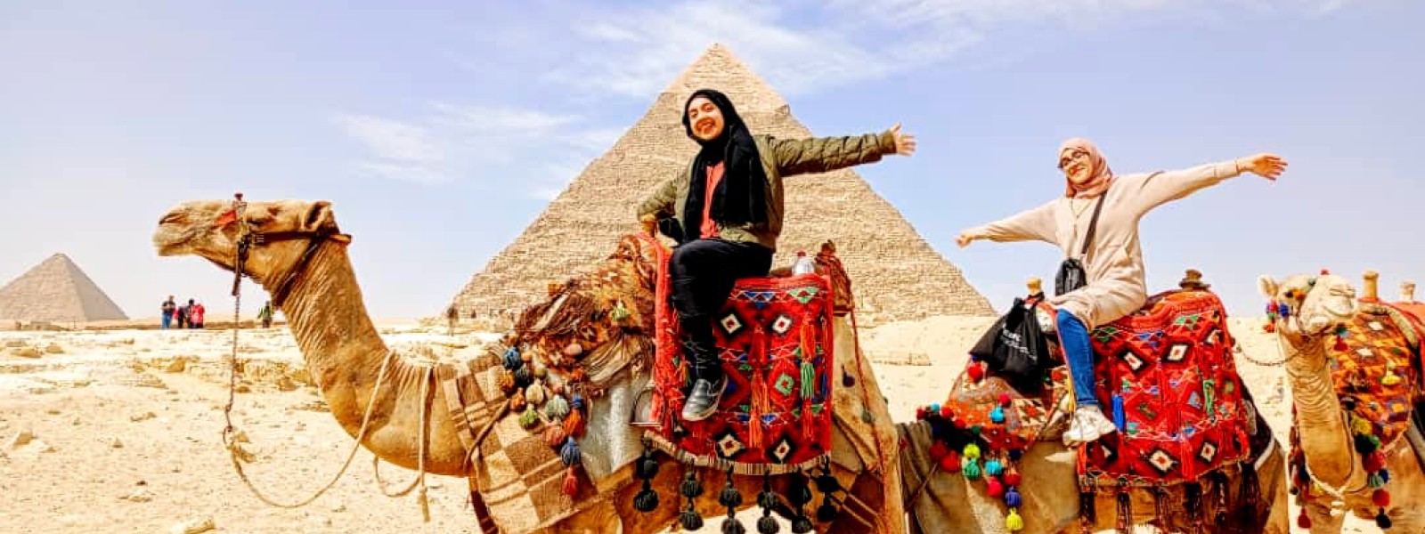Two students on camels pose for a photo. The pyramids can be seen behind them.