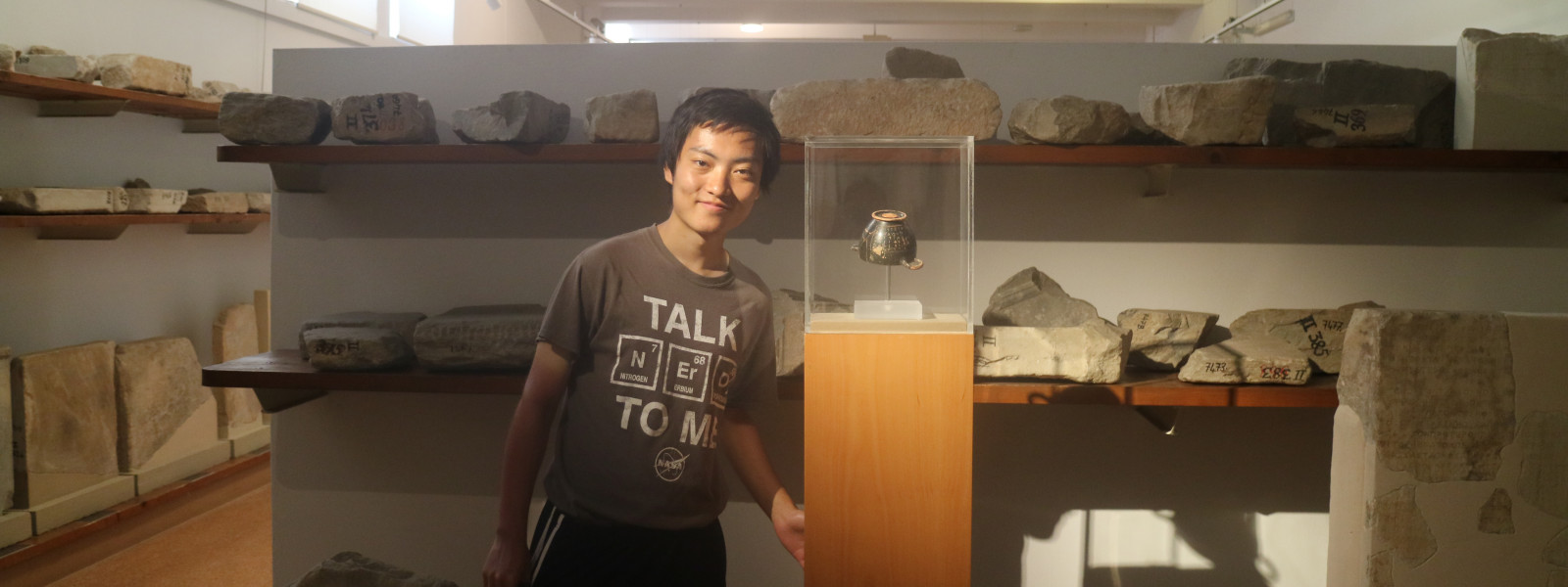 The student is standing next to a display case and there are shelves of artifacts behind him.