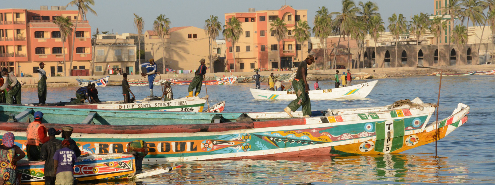 People are walking on and standing beside brightly painted boats by the shore.