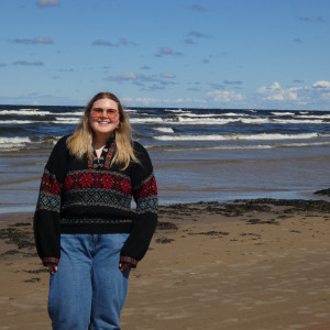 Emma is standing on a beach in a sweater on a sunny day.