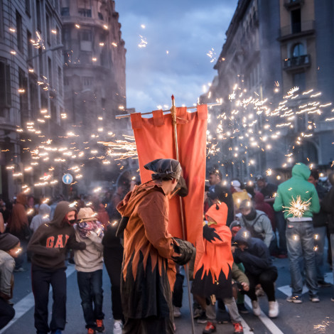 A person wearing a flame-patterned hoodie holds up a gonfalon against a shower of sparks on a crowded street at dusk.