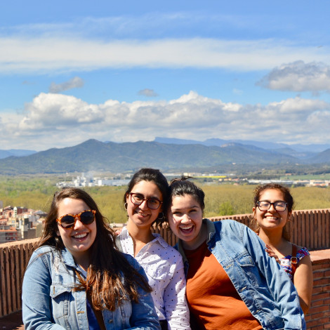 Four students pose for a photo on a sunny day, with buildings, mountains, and clouds behind them.