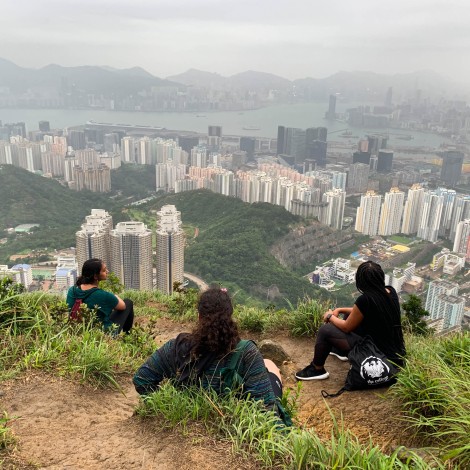 Three students sit and admire the view of buildings, water, and mountains.