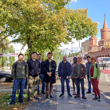 The group stands outside in the shade of a tree before their walking tour. An elevated track and train, cars, and bicyclists can be seen in the background.