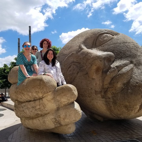 Four students interact with an outdoor sculpture in Paris on a sunny day. They are sitting on or standing beside a giant cupped hand, next to a giant head.
