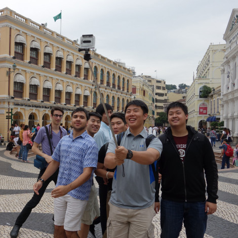 A cluster of smiling students with a camera on a selfie stick are recording video as they explore their surroundings.