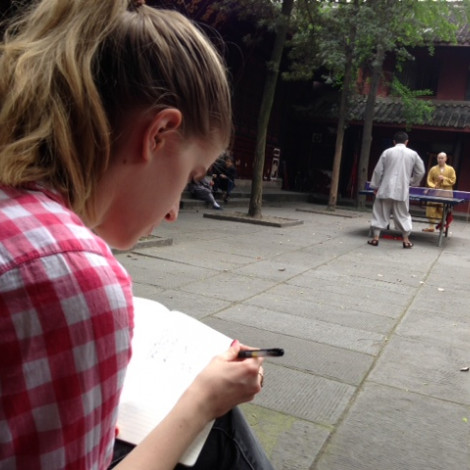 A student is sitting in a courtyard and looking down at a notebook with pen in hand. Two monks play ping pong in the background.